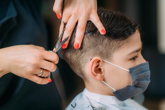 Cortando o cabelo do menino no salão de beleza usando máscara protetora Closeup