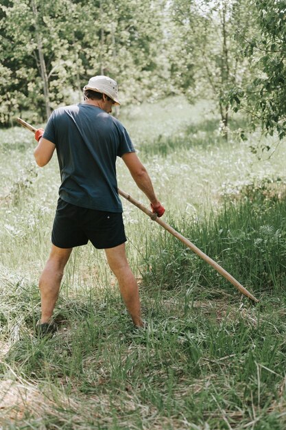 Cortar a grama da maneira tradicional e antiquada com a foice de mão na fazenda da vila doméstica jovem agricultor maduro cortando a grama verde crescida de uma fazenda com uma foice