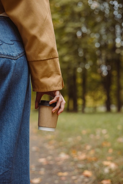 Cortar a mão da mulher em jeans e trincheira segurando uma xícara de papel de café para viagem com espaço de cópia de maquete