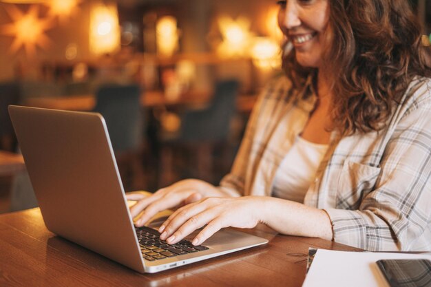 Cortar foto de mulher morena encantadora adulta em camisa xadrez trabalhando com laptop no café