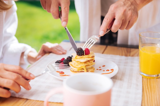Cortar panquecas. Marido carinhoso e amoroso cortando panquecas com frutas e mel em cima para a esposa tomando café da manhã no terraço