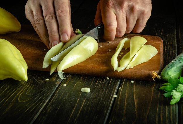 Cortar pimenta para um prato de legumes com uma faca nas mãos de um cozinheiro O conceito de cozinhar dieta vegetal Espaço escuro para receita ou menu