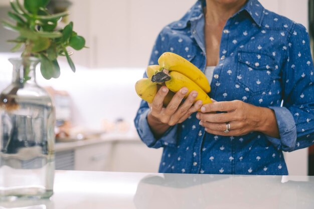 Cortar el retrato de la cara con una mujer en la cocina y la fruta de los plátanos Concepto de personas irreconocibles que trabajan en casa con comida preparando el almuerzo Persona femenina de estilo de vida saludable real con camisa azul