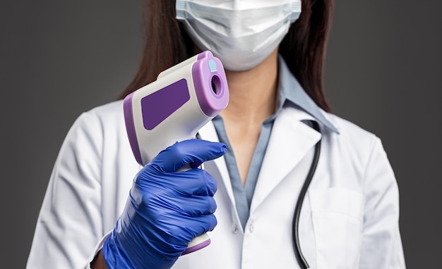 Corte a enfermeira anônima com máscara médica e luvas de proteção usando um moderno termômetro infravermelho sem contato enquanto verifica a temperatura do paciente