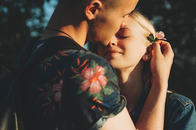 Corte a imagem de um homem atraente anexando ternamente uma delicada flor rosa ao cabelo atrás da orelha de uma adorável jovem loira o abraçando com os olhos fechados