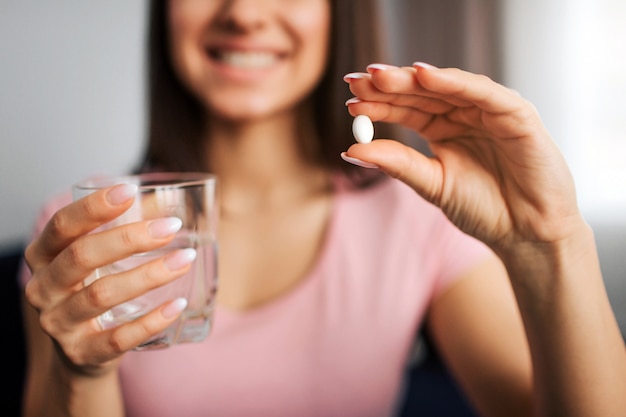 Corte a vista da jovem mulher positiva segura o comprimido branco e o copo de água. Ela sorri para a câmera.