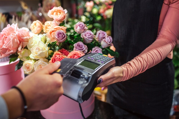 Corte a vista do homem segurando o cartão de crédito acima de dinheiro therminal. Ele paga por flores. Jovem florista feminina segurar dinheiro terminal. Muitas flores estão atrás dela.