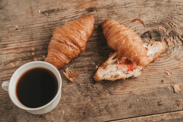 Corte croissant con mantequilla y mermelada de fresa con una taza de café en una vista superior de fondo de madera