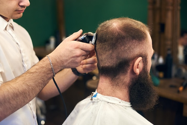 Corte de cabelo masculino profissional para cliente barbudo na barbearia jovem barbeiro trabalhando com barbeador elétrico fechar