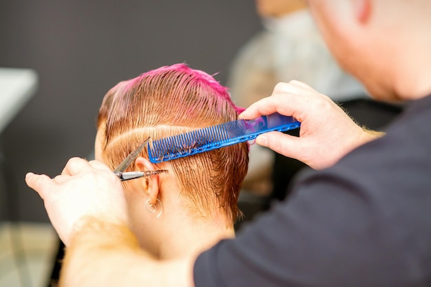 Corte de cabelo tingido de cabelo molhado rosa curto de jovem caucasiana por um cabeleireiro masculino em uma barbearia