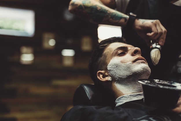 Corte elegante aplicação de espuma nas bochechas do cliente para barbear enquanto trabalha na barbearia