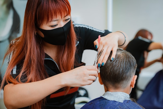 Foto corte de pelo al niño al cliente con medidas de seguridad. estilista asiático. reanudación del trabajo con medidas de seguridad de barbería en el contexto de la pandemia covid-19