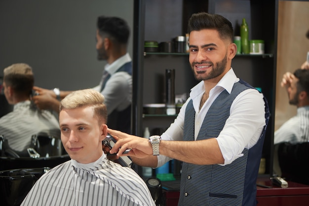 Corte de pelo del cliente de corte de peluquero guapo, mirando a cámara y sonriendo.