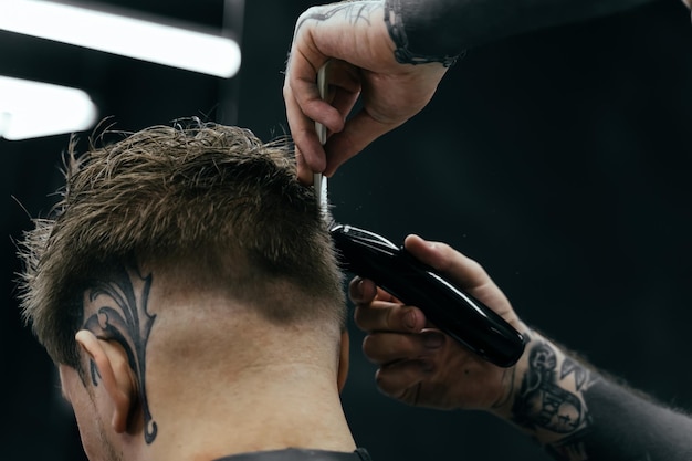 Corte de pelo masculino con maquinilla de afeitar eléctrica Peluquero tatuado hace corte de pelo para el cliente en la barbería usando cortapelos Hombre peluquería con maquinilla de afeitar eléctrica