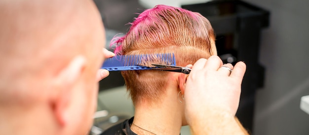 Corte de pelo de pelo húmedo teñido de rosa corto de una joven caucásica por un peluquero masculino en una barbería