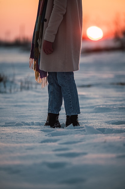 Corte uma mulher irreconhecível com roupas quentes e botas na neve branca fresca na zona rural de inverno ao pôr do sol