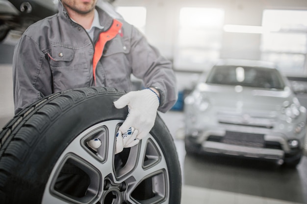 Corte la vista del trabajador en un uniforme gris y sostenga la rueda del automóvil con ambas manos.