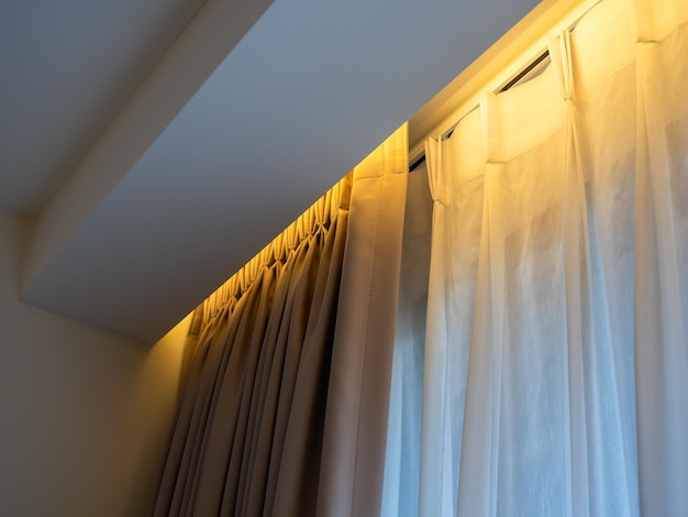 Cortina opaca beige y cortinas de lino blanco translúcido en el riel de la cortina decoradas con luz amarilla escondida bajo el techo cerca de la ventana corrediza de vidrio en el dormitorio blanco