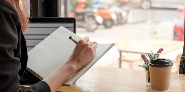 Cosecha cerca del estudio de la mujer en línea distante en la computadora portátil hacer nota escribir en el cuaderno sentado en el escritorio en casa. Escritora inteligente o estudiante escribe a mano en el bloc de notas de papelería, planificación o lista de elaboración.