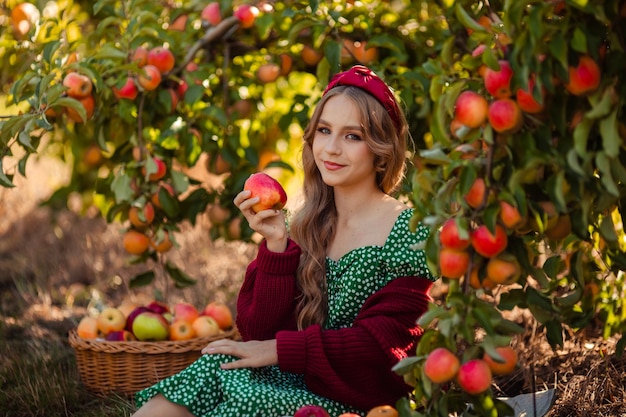 Cosechando en el retrato de la granja de una hermosa chica con un vestido verde en un huerto de manzanas con una cesta