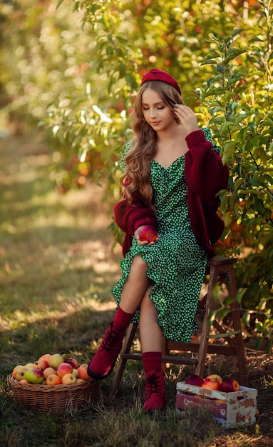 Cosechando en el retrato de la granja de una hermosa chica con un vestido verde en un huerto de manzanas con una cesta