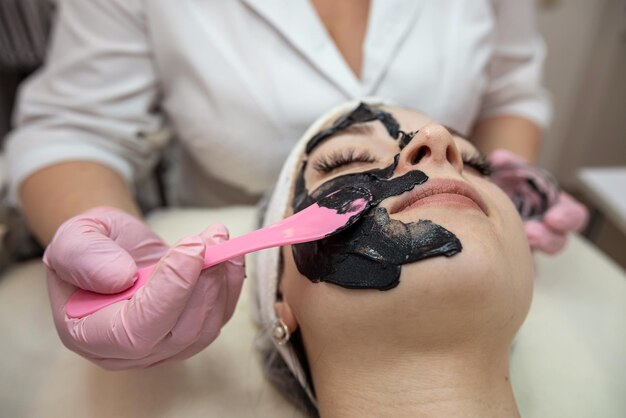 Cosmética aplicando máscara preta no rosto de uma jovem e bonita mulher no salão de spa Conceito de procedimento cosmético anti-envelhecimento