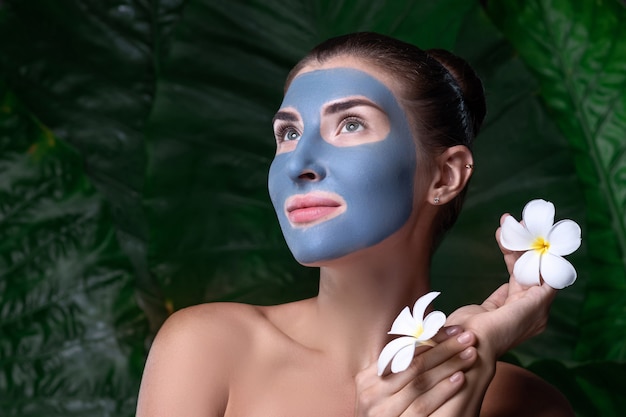 Cosmética ecológica. Una mujer pensativa con una máscara de arcilla azul en su rostro y con flores blancas en sus manos