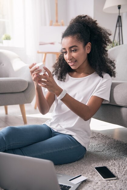 Cosmética facial. Mujer joven positiva mirando la crema facial mientras está cerca de su computadora portátil