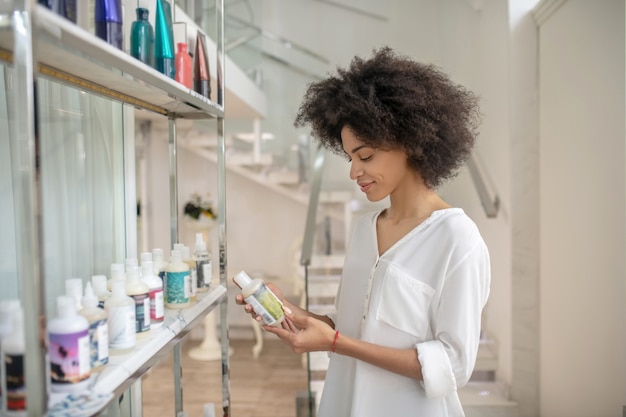 Cosméticos de cuidado. Niña bonita sonriente joven interesada en productos cosméticos de pie en el vestíbulo del centro de belleza