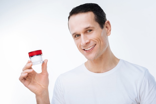 Cosméticos faciais. Homem simpático e positivo sorrindo e segurando uma garrafa de creme facial enquanto o usa