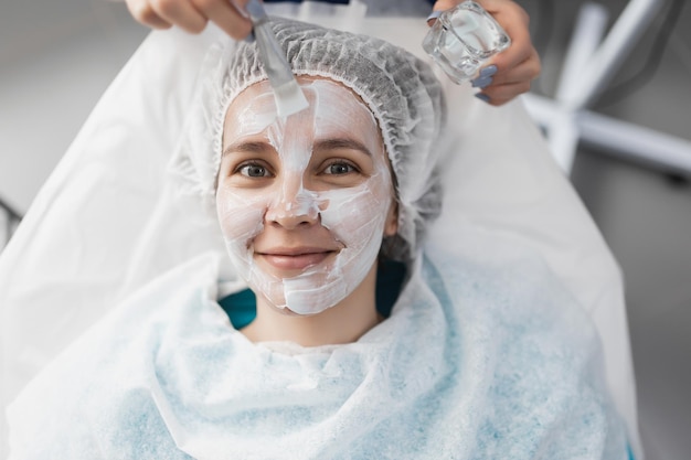 La cosmetóloga aplica una máscara de crema blanca hidratante en la cara a un paciente sonriente con un cepillo