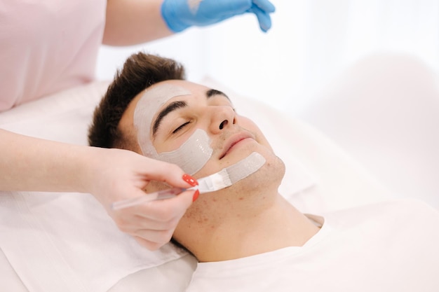 Cosmetóloga aplicando máscara de arcilla en la cara del hombre en el salón de spa primer plano de la cara masculina durante el procedimiento