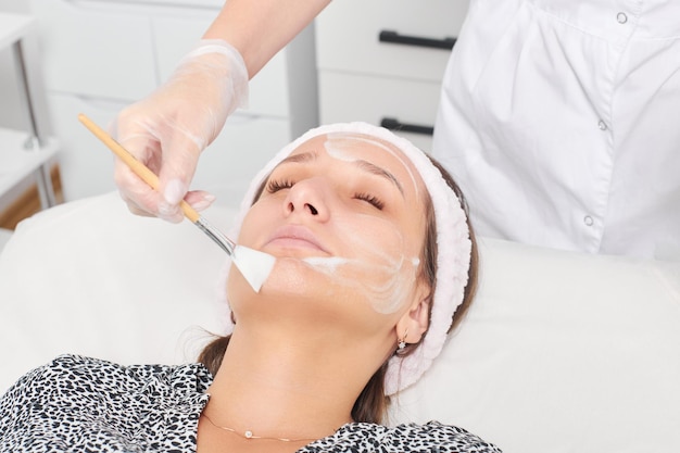 Cosmetóloga aplicando máscara de crema cosmética en la cara femenina para el procedimiento de rejuvenecimiento en el salón de belleza