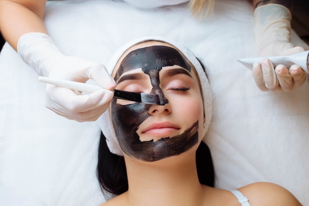 Cosmetóloga aplicando máscara negra en la cara de una mujer bonita con guantes negros hermosa mujer en spa