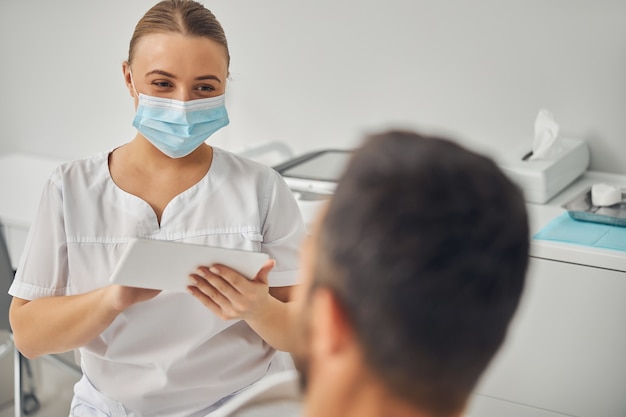 Cosmetóloga femenina en mascarilla médica con almohadilla electrónica PC mientras conversa con el joven