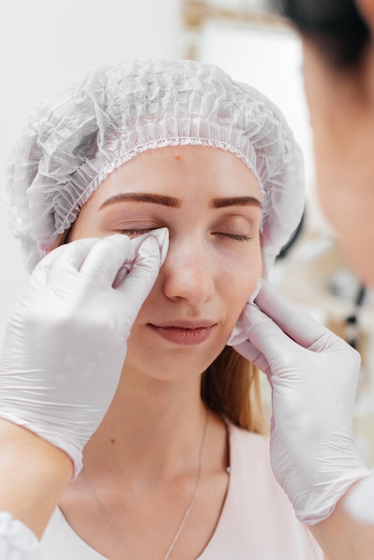 La cosmetóloga limpia la cara del paciente después del procedimiento de llenado del surco nasolagrimal y mesoterapia alrededor de los ojos para una joven hermosa Cosmetología moderna