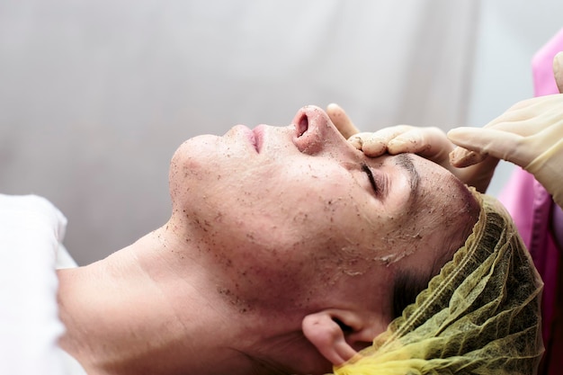La cosmetóloga limpia la piel de la cara del paciente con un exfoliante en la oficina.