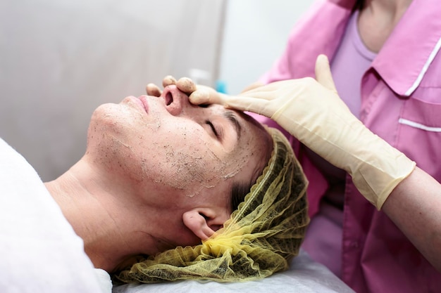 La cosmetóloga limpia la piel de la cara del paciente con un exfoliante en la oficina.