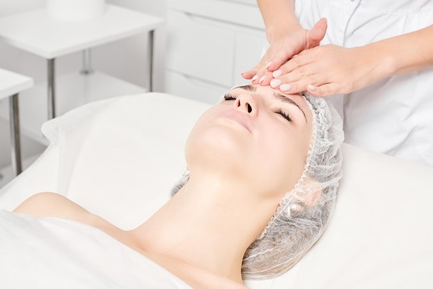 La cosmetóloga masajea la máscara de crema en la piel de la cara de la mujer para el procedimiento de rejuvenecimiento en el salón de belleza