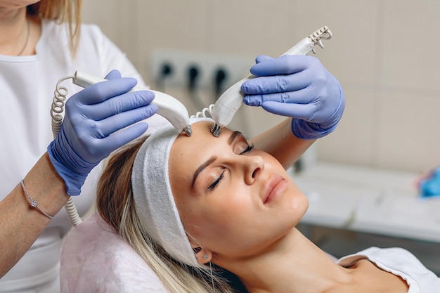 Cosmetología, adorable encantadora joven en un salón de spa disfrutando de un estimulante tratamiento facial eléctrico.