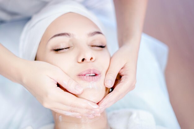 Cosmetologia As mãos de um cosmetologista fazem uma massagem facial com uma máscara Jovem linda no procedimento de spa