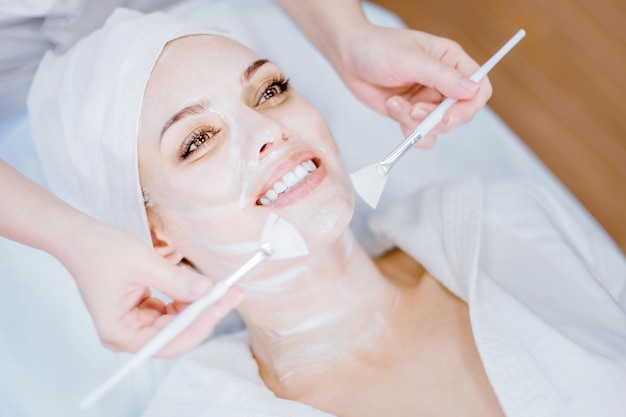 Cosmetología La cosmetóloga aplica una mascarilla facial limpiadora con dos cepillos Niña sonriente en el procedimiento de rejuvenecimiento facial Procedimiento de spa