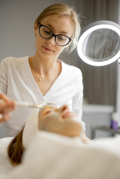 Foto cosmetologista aplica máscara de beleza com pincel no rosto de uma mulher no salão de beleza conceito de cuidados faciais e procedimentos de beleza