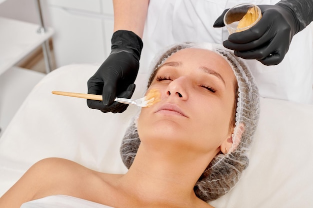 Cosmetologista aplica máscara de mel no rosto da mulher para hidratar a pele do rosto no salão de spa de beleza