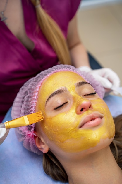 Cosmetologista aplica máscara dourada com pincel no rosto da mulher. pele facial e tratamento anti-envelhecimento. cosmetologia e cuidados profissionais da pele facial.