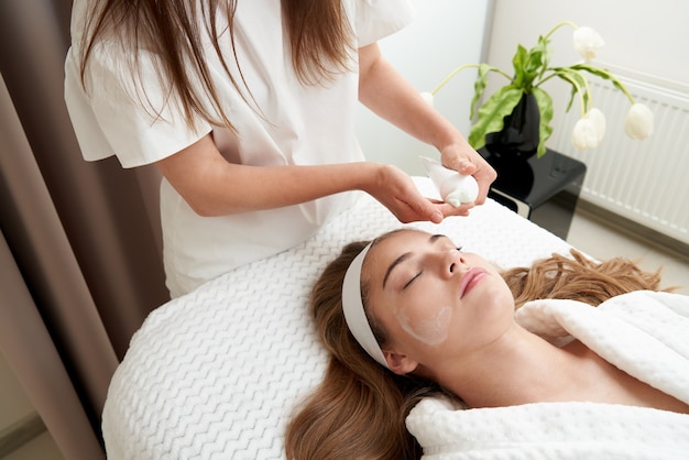 Cosmetologista aplicando creme no rosto feminino de mulher fazendo tratamento facial em salão de spa