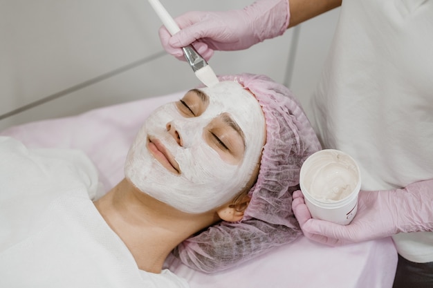 Cosmetologista aplicando máscara no rosto do cliente em salão de spa jovem recebendo cuidados faciais