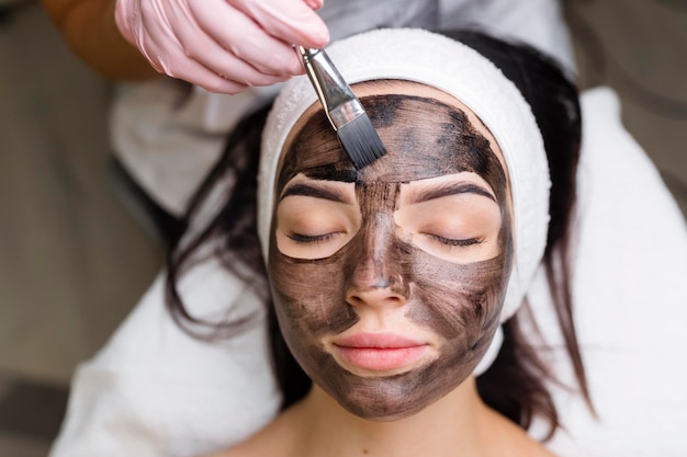 Cosmetologista aplicando máscara preta no rosto de mulher bonita usando luvas pretas linda mulher no spa tendo procedimentos faciais