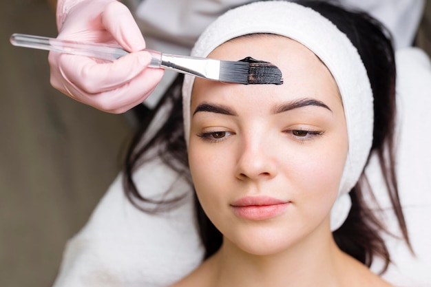 Cosmetologista aplicando máscara preta no rosto de mulher bonita usando luvas pretas linda mulher no spa tendo procedimentos faciais