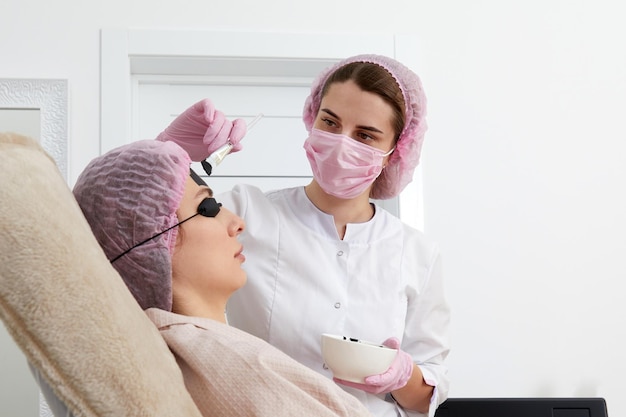 Cosmetologista aplicando máscara preta no rosto de uma linda mulher para peeling de carbono